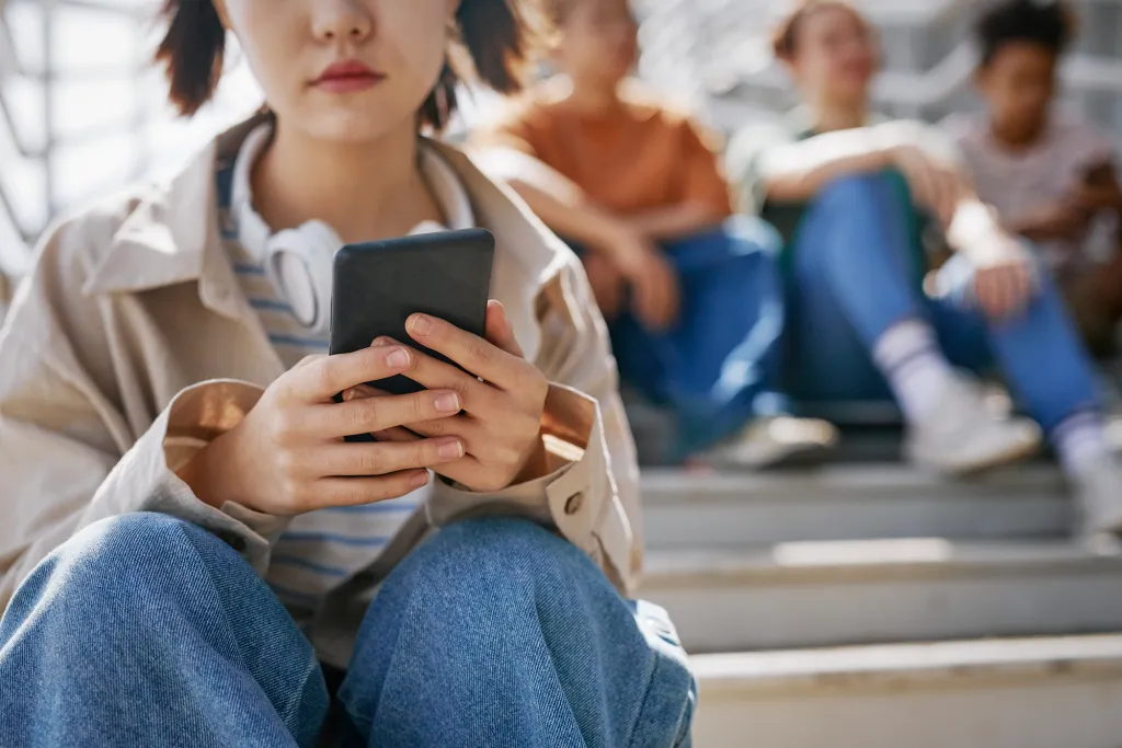 A teenager holds a smartphone while sitting on stairs with a group of friends. Photo via iStock